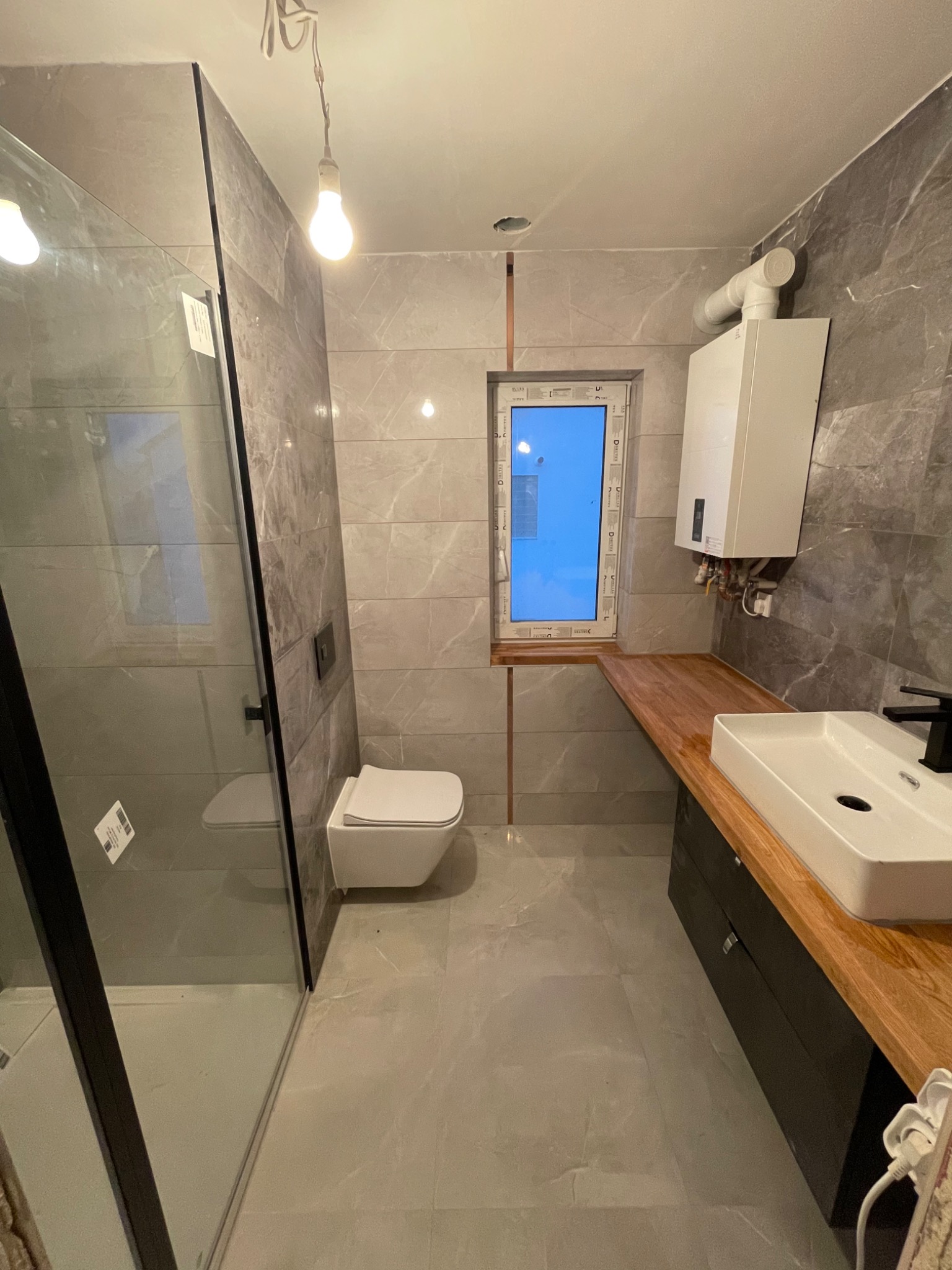Nowoczesna łazienka z szarymi płytkami imitującymi marmur, kabiną prysznicową typu walk-in z czarnymi profilami, wiszącą toaletą, drewnianym blatem pod umywalką nablatową i piecem gazowym na ścianie.