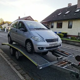 Srebrny Mercedes Klasy A na lawecie samochodowej, widok z przodu na tle domów jednorodzinnych.