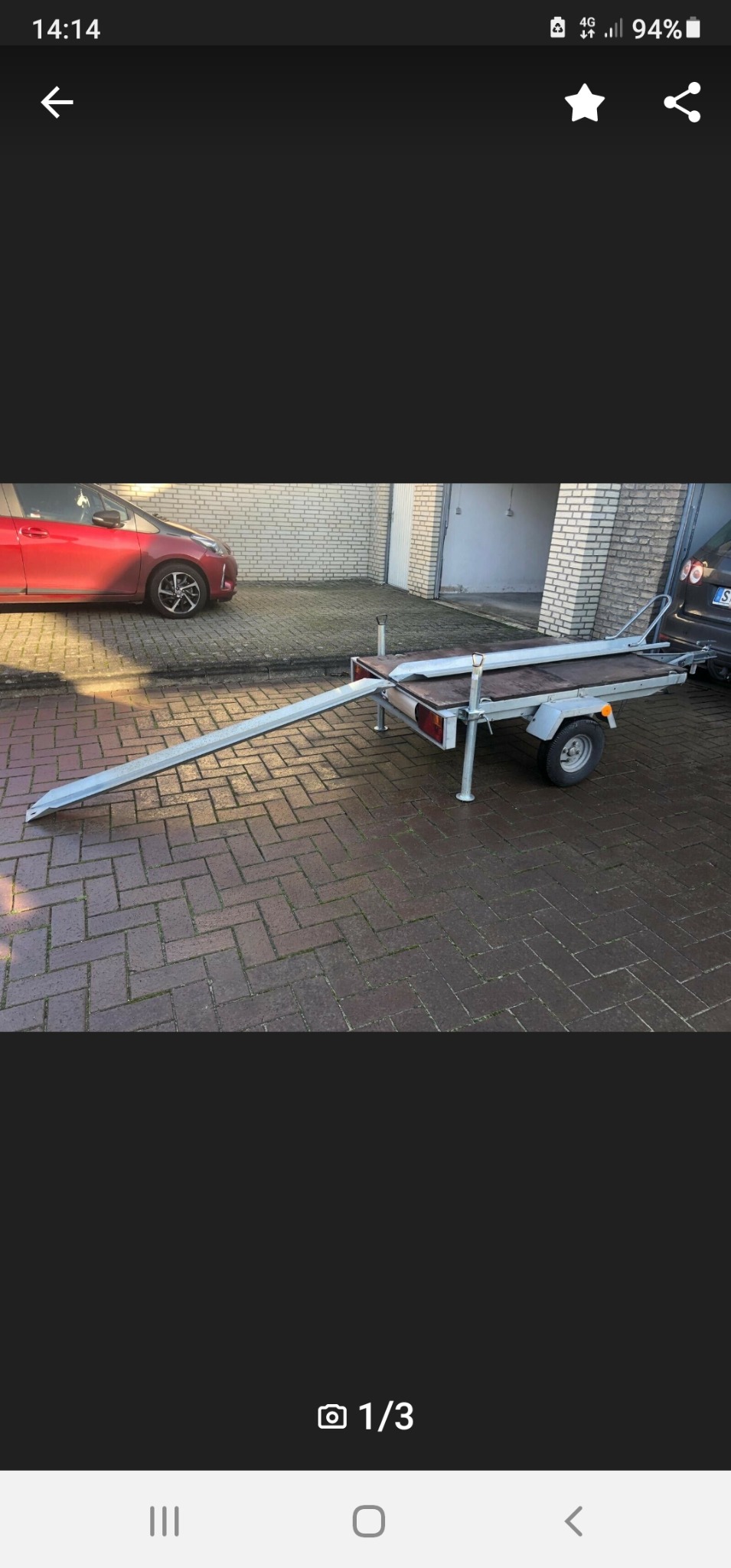 Mała przyczepka samochodowa z najazdem, zaparkowana na brukowanym podjeździe przed garażem i czerwonym samochodem.