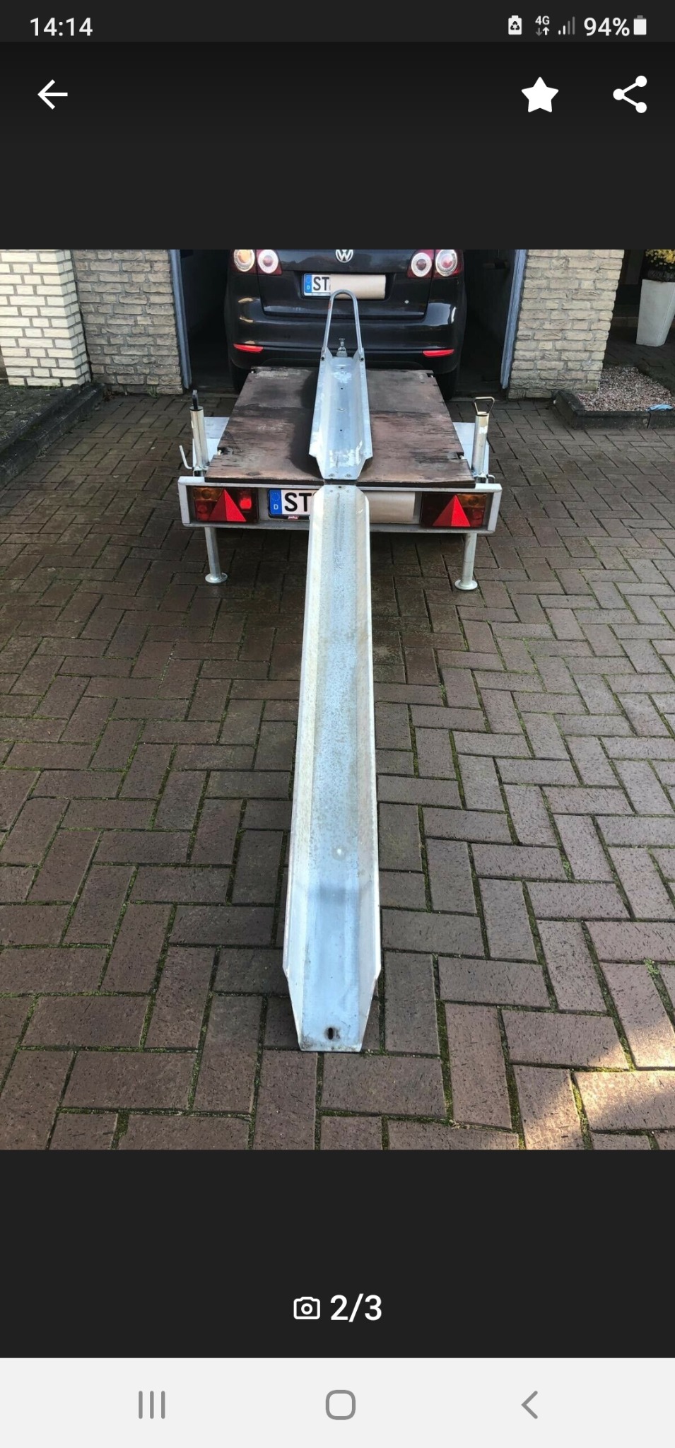 Rampa najazdowa do przyczepy samochodowej ustawiona na kostce brukowej, z widocznym fragmentem przyczepy i zaparkowanym samochodem Volkswagen w garażu.