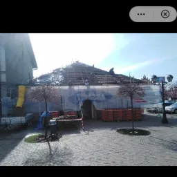 Renowacja dachu budynku w centrum miasta, widoczne rusztowania, materiały budowlane i ekipa remontowa pracująca na dachu.