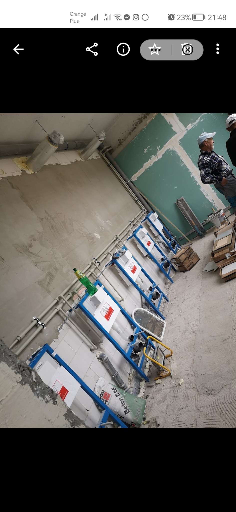 Wnętrze łazienki w trakcie remontu, widoczne stelaże podtynkowe WC z niebieską ramą, instalacje hydrauliczne, surowa ściana z płyt kartonowo-gipsowych i pracownik w tle.