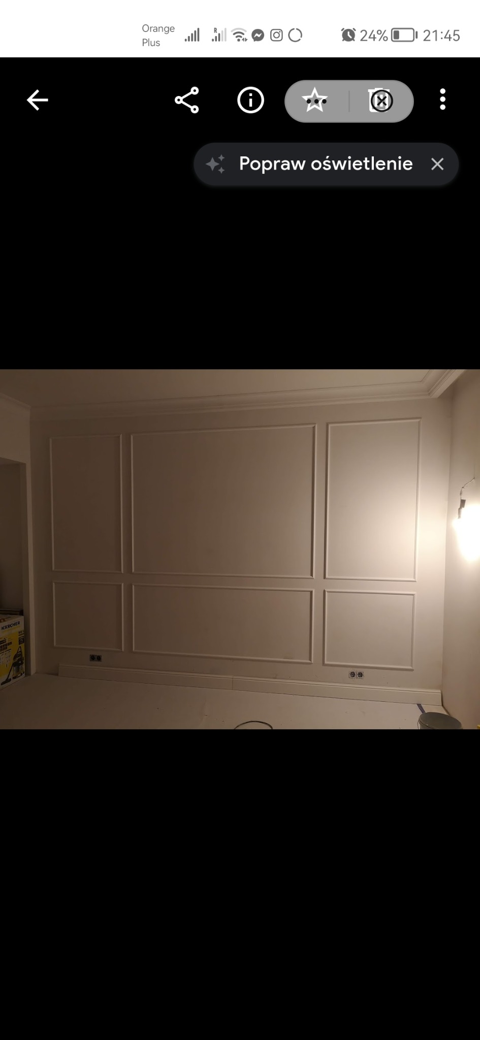 Ściana wewnętrzna z dekoracyjnymi panelami w stylu klasycznym, pomalowana na jasny kolor, widoczne gniazdka elektryczne i listwa przypodłogowa, w tle częściowo widoczny odkurzacz Karcher.