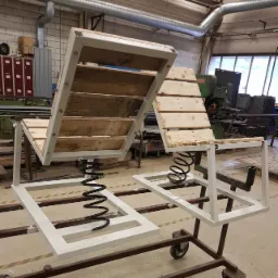 Dwa unikalne krzesła ogrodowe na sprężynach, z metalową ramą pomalowaną na biało i siedziskami z desek, umieszczone na wózku w warsztacie.