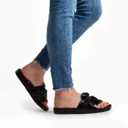 Stopa w dżinsach i czarnych lakierowanych klapkach z kokardą na białym tle.