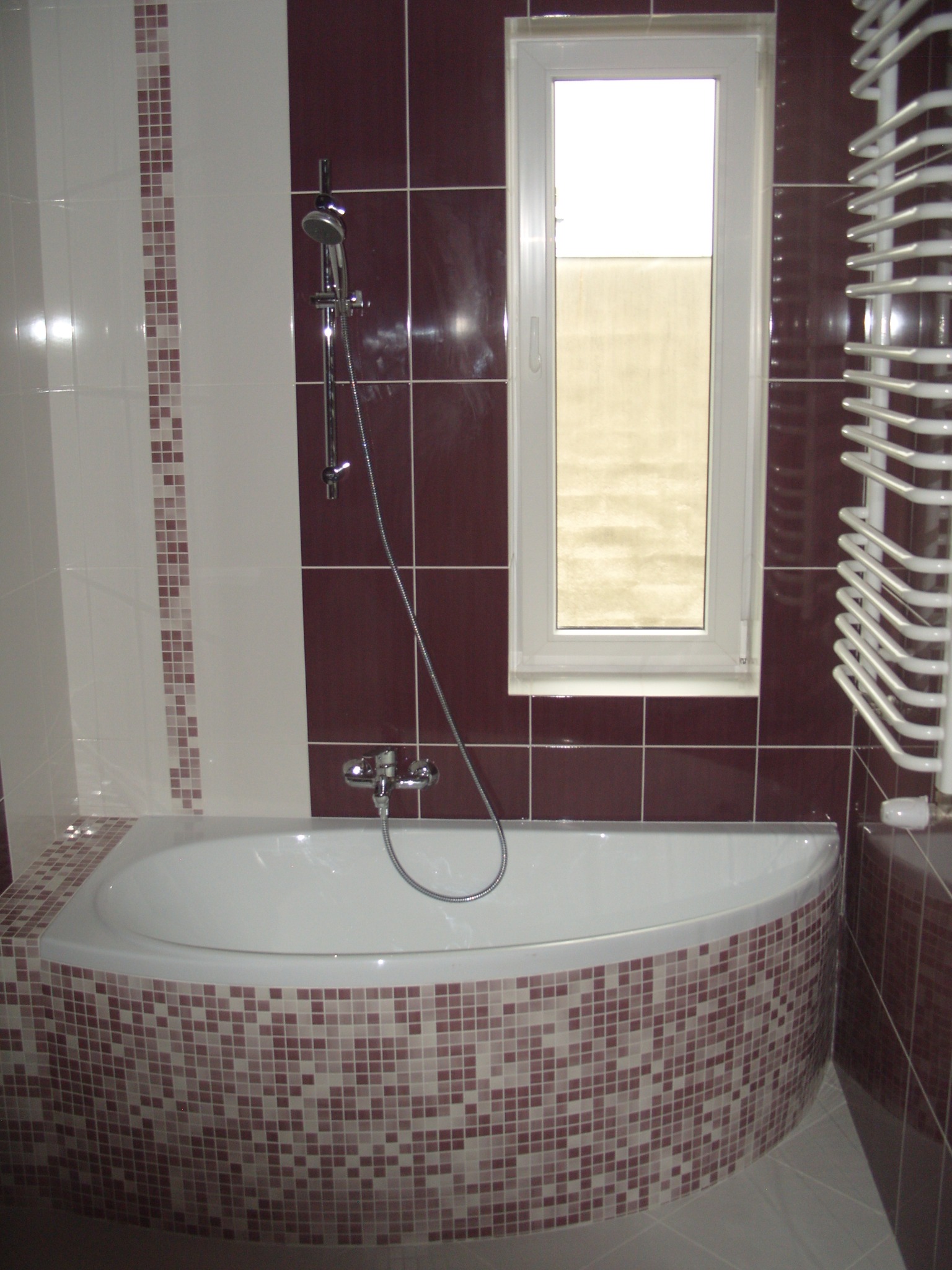 Biała wanna narożna obłożona mozaiką w odcieniach brązu i beżu, dopasowana do ciemnobrązowych płytek ściennych i białych paneli z mozaikowym paskiem, z widocznym grzejnikiem drabinkowym i oknem w tle.