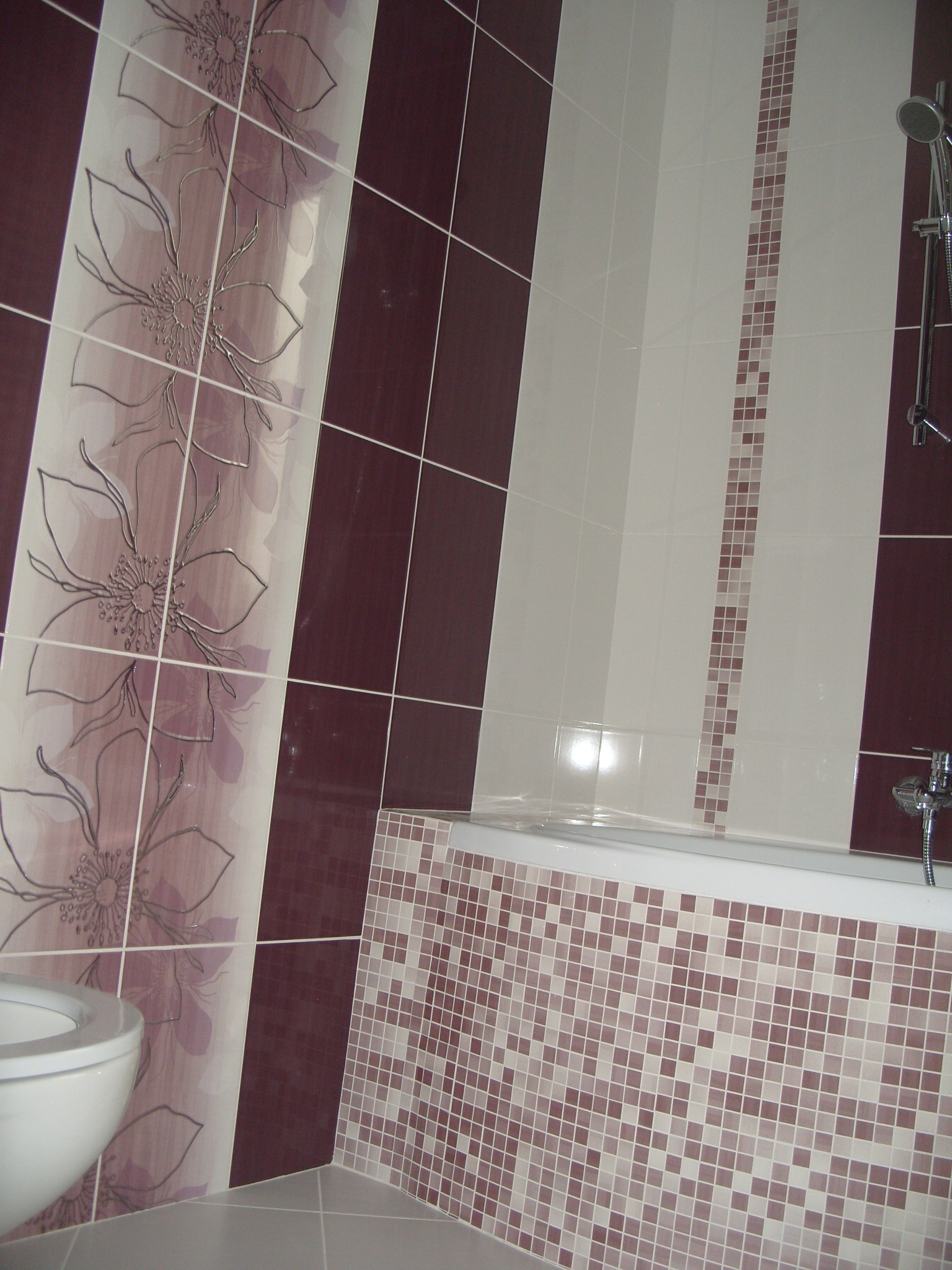 Ukończona łazienka z bordowo-białymi płytkami, dekoracyjnym motywem kwiatowym na ścianie, mozaiką na obudowie wanny i widoczną toaletą.