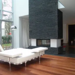 Nowoczesny salon z drewnianą podłogą, białym leżakiem, kamiennym kominkiem i dużym oknem z widokiem na zieleń.