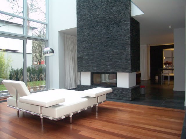 Nowoczesny salon z drewnianą podłogą, białym leżakiem, kamiennym kominkiem i dużym oknem z widokiem na zieleń.