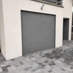 Szara, ziarnista elewacja garażu przylegającego do jasnego budynku, oświetlona przez pojedynczy kinkiet, z kostką brukową na podjeździe.