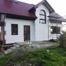 Dom jednorodzinny w trakcie ocieplania elewacji styropianem, widoczne rusztowania i niezabezpieczone krawędzie styropianu, przygotowane schody wejściowe.