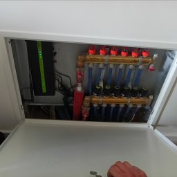 AUTO CZĘŚCI FIREK - MARCIN FIREK - Otwarta szafka z rozdzielaczem ogrzewania podłogowego, widoczne rury, zawory i sterownik. Na górnej belce rozdzielacza widoczne napisy.