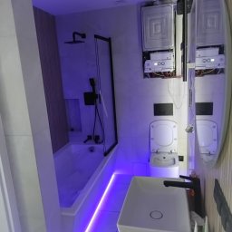 AUTO CZĘŚCI FIREK - MARCIN FIREK - Nowoczesna łazienka z wanną, kabiną prysznicową, umywalką i toaletą w bieli, oświetlona fioletowym światłem LED. Widoczne dwa bojlery na ścianie.