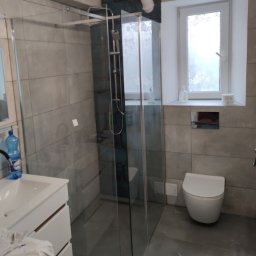 AUTO CZĘŚCI FIREK - MARCIN FIREK - Wykończona łazienka z kabiną prysznicową typu walk-in, umywalką z szafką i toaletą. Szare płytki imitujące beton na ścianach i podłodze. Okno z białymi ramami.