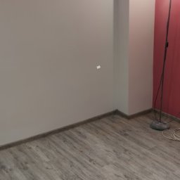 AUTO CZĘŚCI FIREK - MARCIN FIREK - Jasne wnętrze po remoncie: gładkie, szare ściany, drewnopodobna podłoga, pionowy filar i lampa podłogowa przy czerwonej ścianie. Minimalistyczny design.