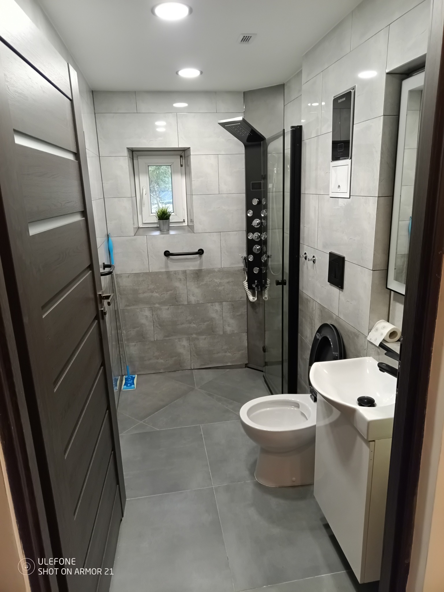 Nowoczesna łazienka z szarymi płytkami, czarną armaturą i kabiną prysznicową typu walk-in, widok z perspektywy drzwi wejściowych.