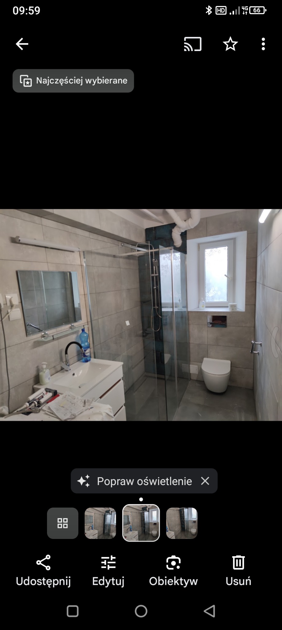 Wyremontowana łazienka z kabiną prysznicową, umywalką z szafką, toaletą i szarymi płytkami na ścianach. Na umywalce widoczne akcesoria łazienkowe oraz narzędzia.