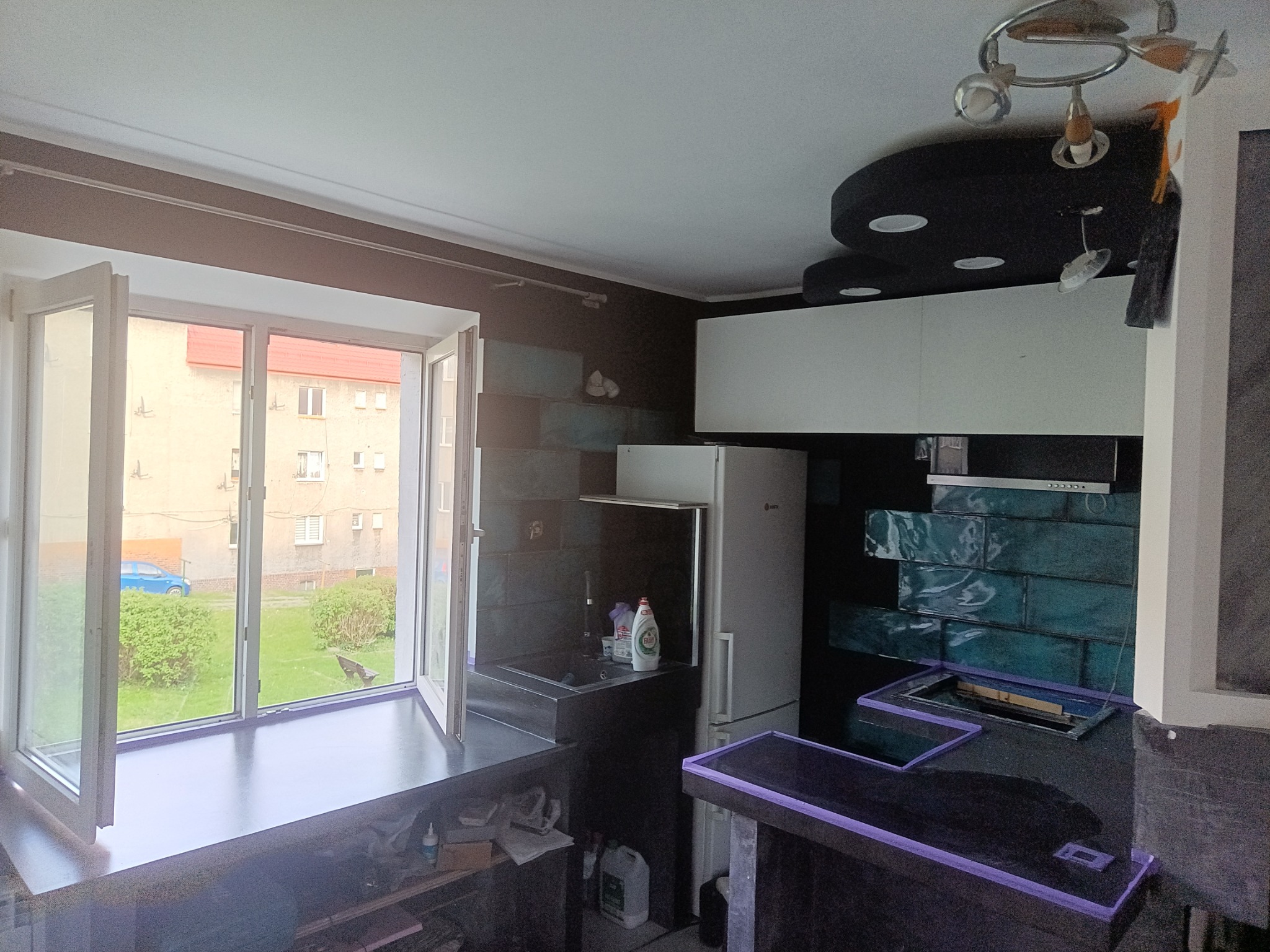 Ciemna kuchnia z turkusowymi płytkami na ścianie, widok na otwarte okno z zielenią na zewnątrz, fragment sufitu z designerskim oświetleniem.