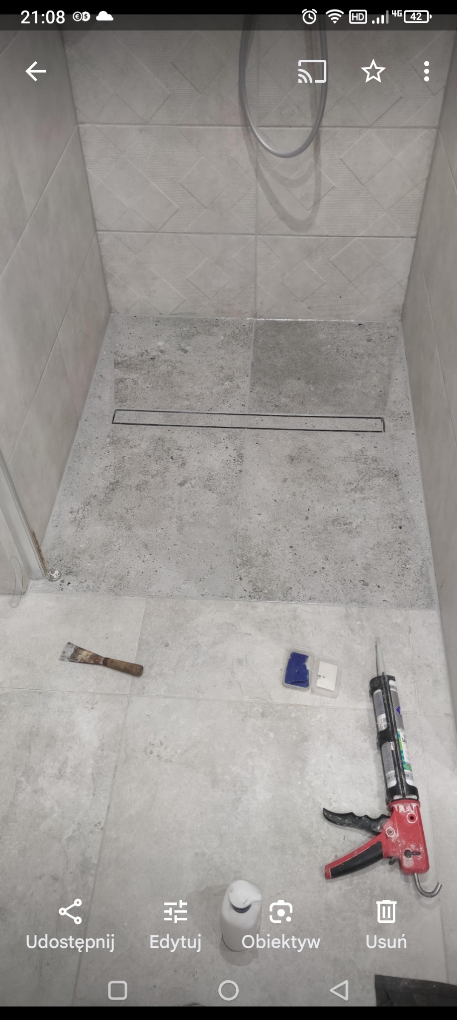 Wykończony brodzik z odpływem liniowym, widoczne płytki imitujące beton, narzędzia glazurnicze: szpachelka, fuga w opakowaniu i pistolet do silikonu.