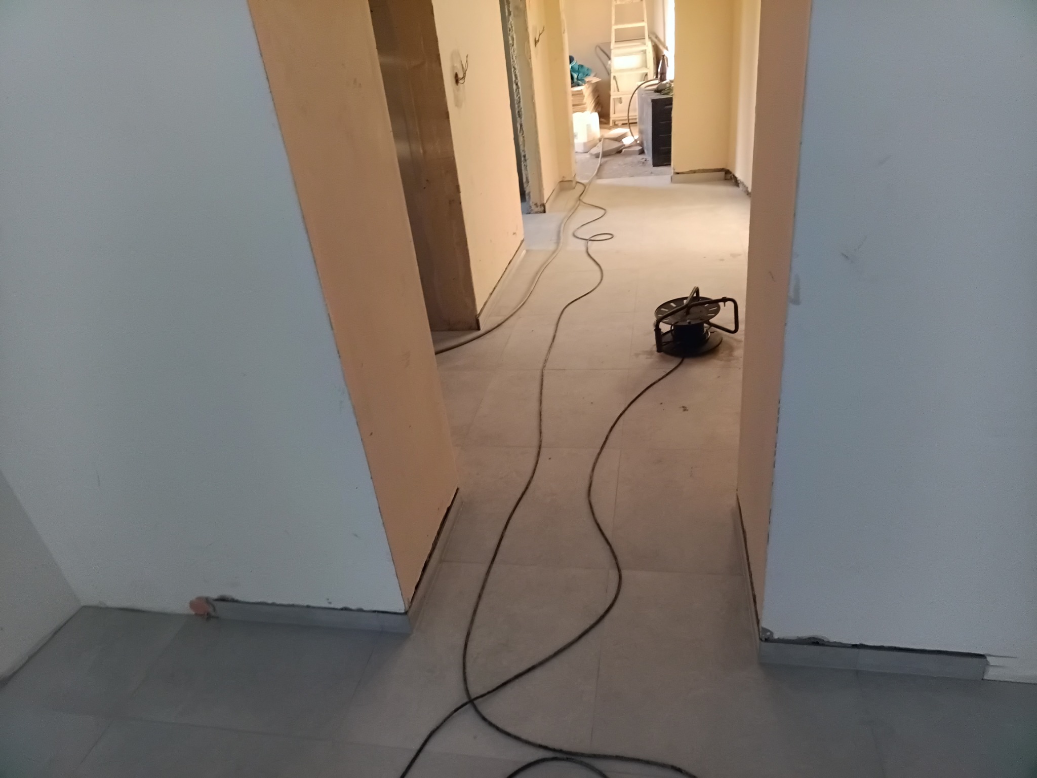 Korytarz w trakcie prac wykończeniowych, widoczne ściany przygotowane do malowania, szare płytki na podłodze oraz przewód elektryczny z przedłużaczem. W głębi pomieszczenia drabina i materiały...