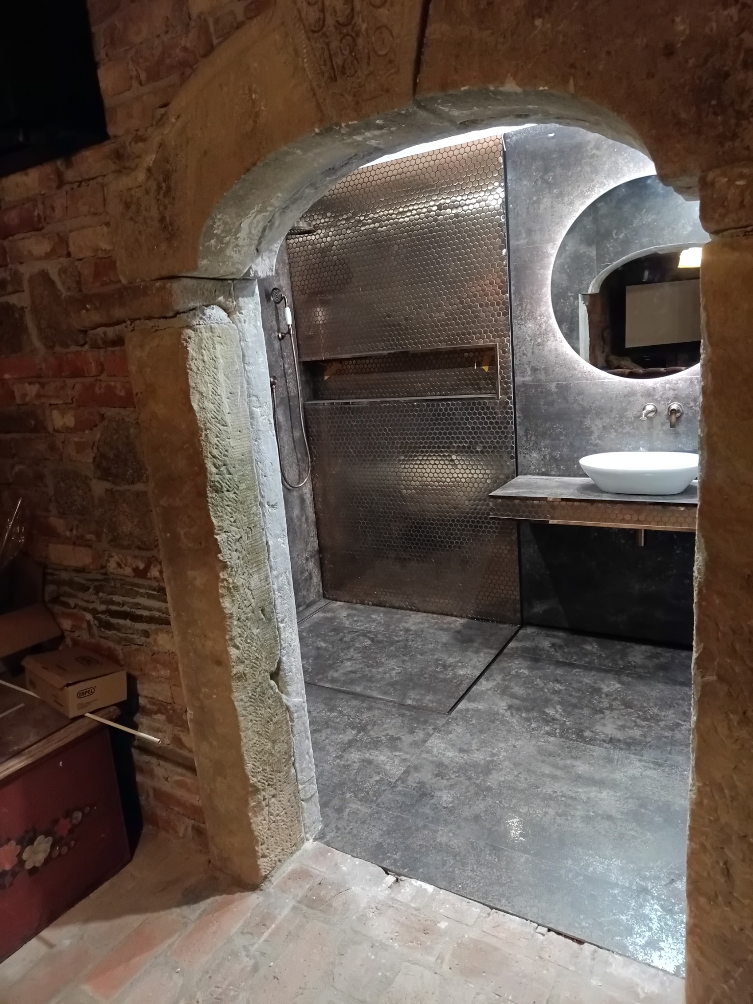 Nowoczesna łazienka z kabiną prysznicową wykończoną metalową mozaiką, umywalką na blacie i okrągłym lustrem z podświetleniem, widoczna przez kamienny łuk.