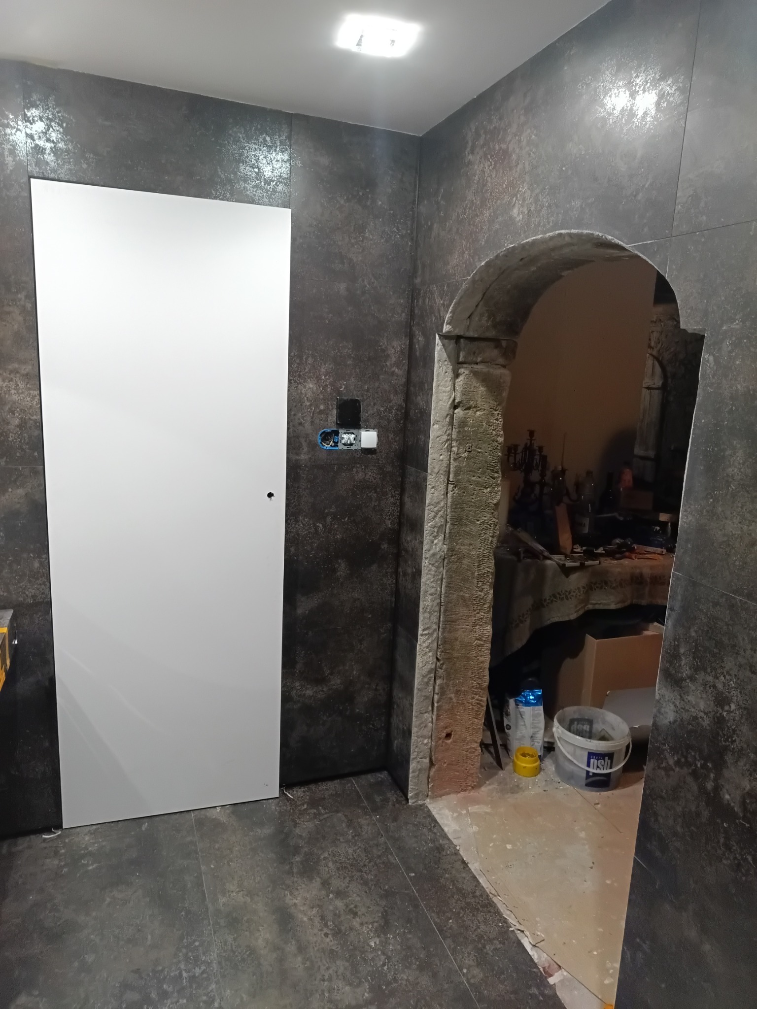 Wnętrze łazienki z szarą, strukturalną okładziną ścienną, białe drzwi po lewej stronie z widoczną instalacją elektryczną obok, oraz kamienne, łukowe przejście do innego pomieszczenia z widocznymi...