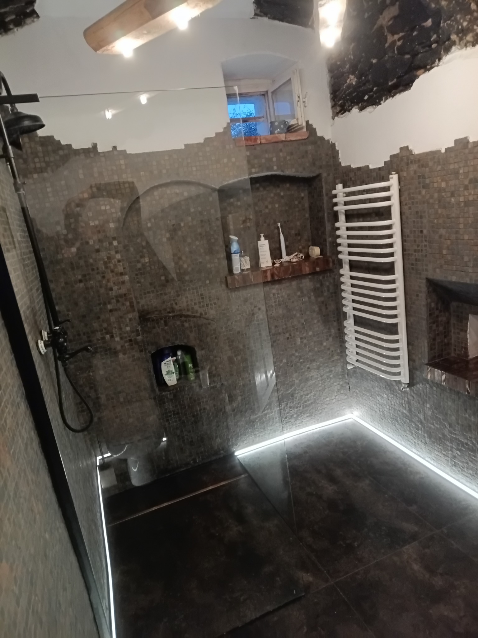 Nowoczesna łazienka z kabiną prysznicową typu walk-in, wykończona mozaiką w odcieniach szarości, z oświetleniem LED przy podłodze i grzejnikiem drabinkowym.