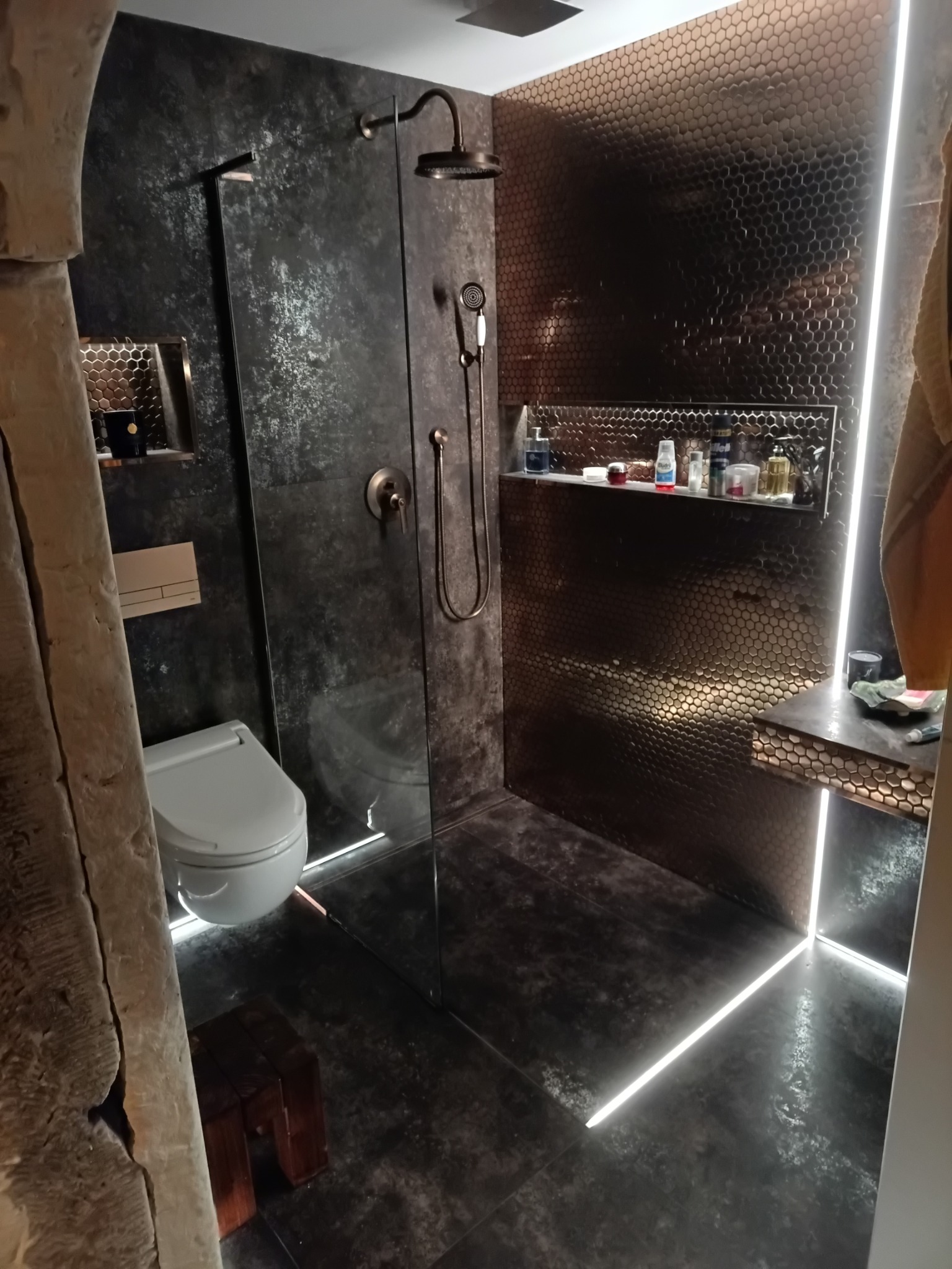 Nowoczesna łazienka z kabiną prysznicową typu walk-in, wykończona ciemnymi płytkami imitującymi kamień oraz mozaiką w odcieniach brązu, z subtelnym oświetleniem LED podkreślającym detale.