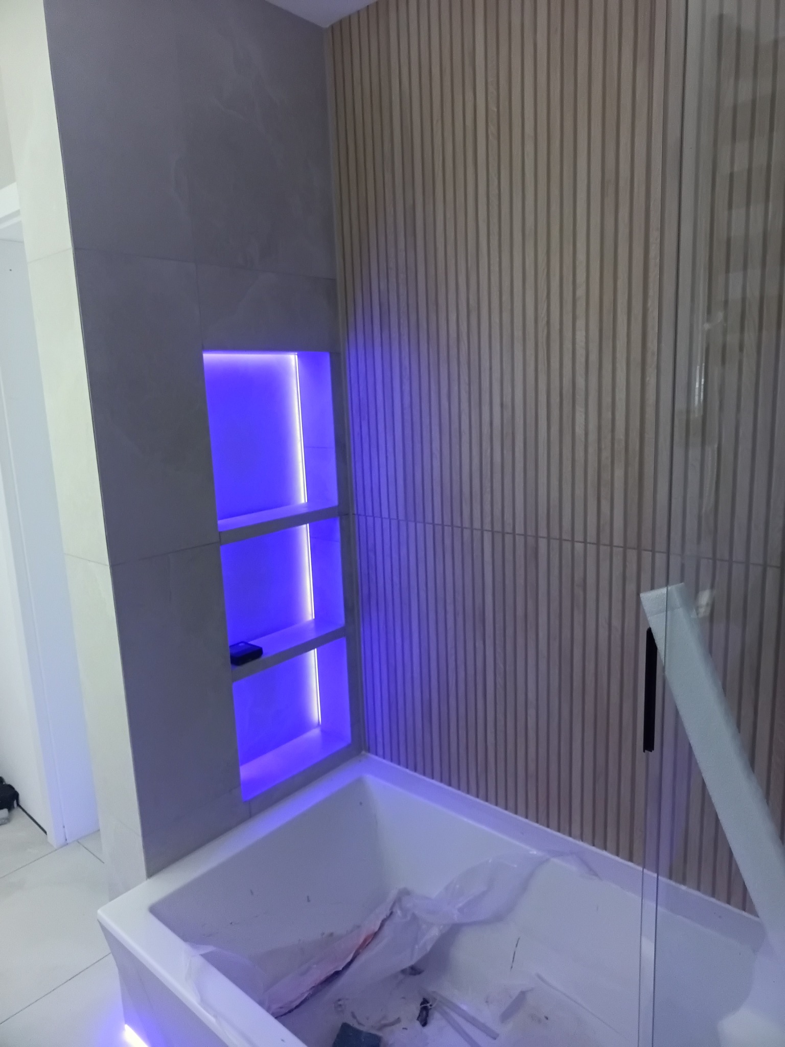 Wykończona łazienka z podświetlaną wnęką w szarej płytce i drewnianą ścianą. Widoczna wanna z elementami montażowymi. Nowoczesny design z akcentem na oświetlenie.