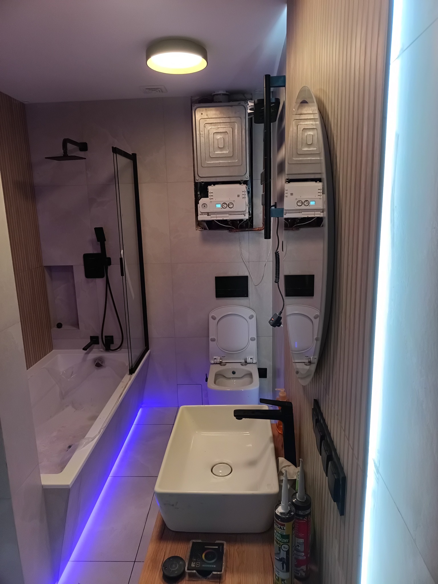 Nowoczesna łazienka z podświetleniem LED, białą armaturą i czarnymi detalami. Uwagę zwraca nietypowe umiejscowienie pieca gazowego nad toaletą. Minimalistyczny design.