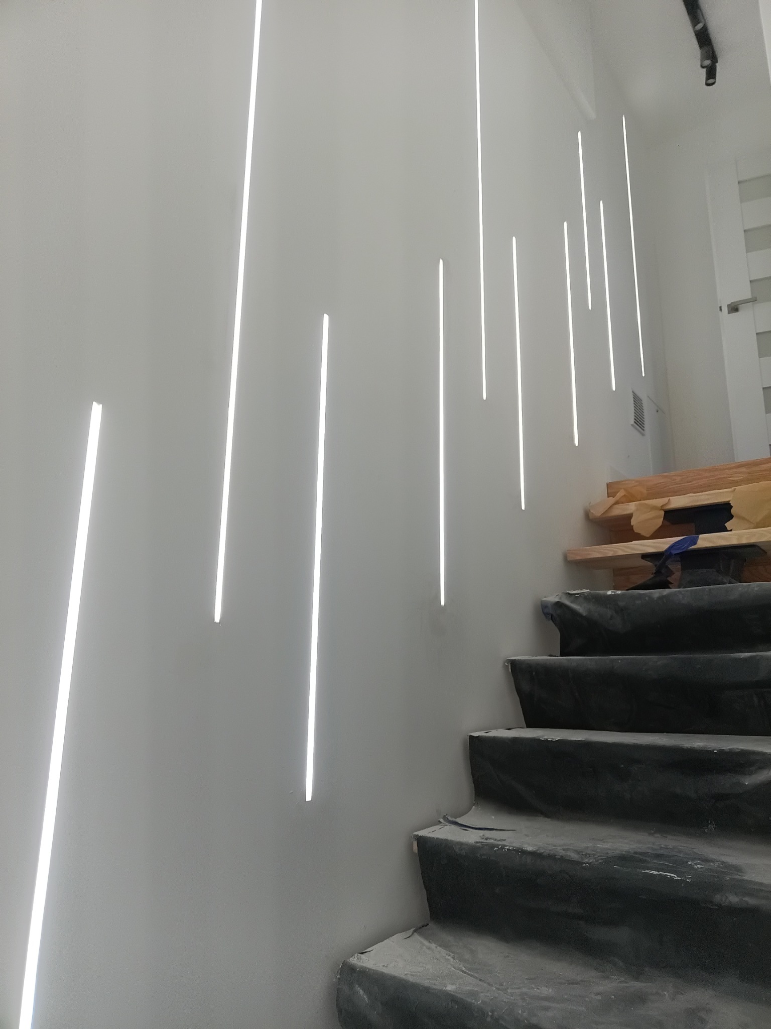 Nowoczesne oświetlenie schodowe: pionowe, wpuszczane listwy LED na białej ścianie, schody w trakcie wykończenia, zabezpieczone folią. Minimalistyczny design.