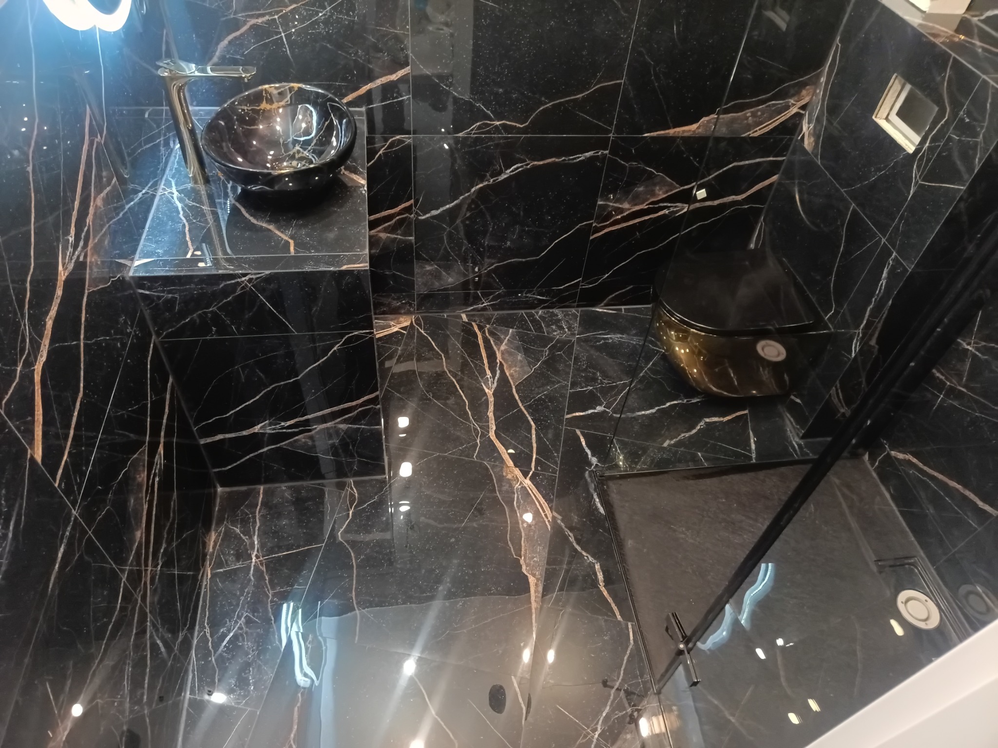 Luksusowa łazienka wykończona czarnym marmurem z złotymi żyłami. Umywalka nablatowa i toaleta w kolorze czarnym, kabina prysznicowa ze szklanymi drzwiami. Nowoczesny design.