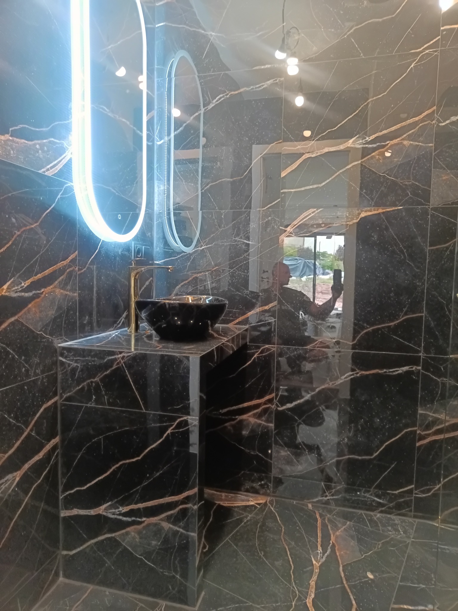 Elegancka łazienka z czarnymi płytkami imitującymi marmur, podświetlane lustro owalne i czarna umywalka nablatowa ze złotą armaturą. Widoczne odbicie osoby robiącej zdjęcie.