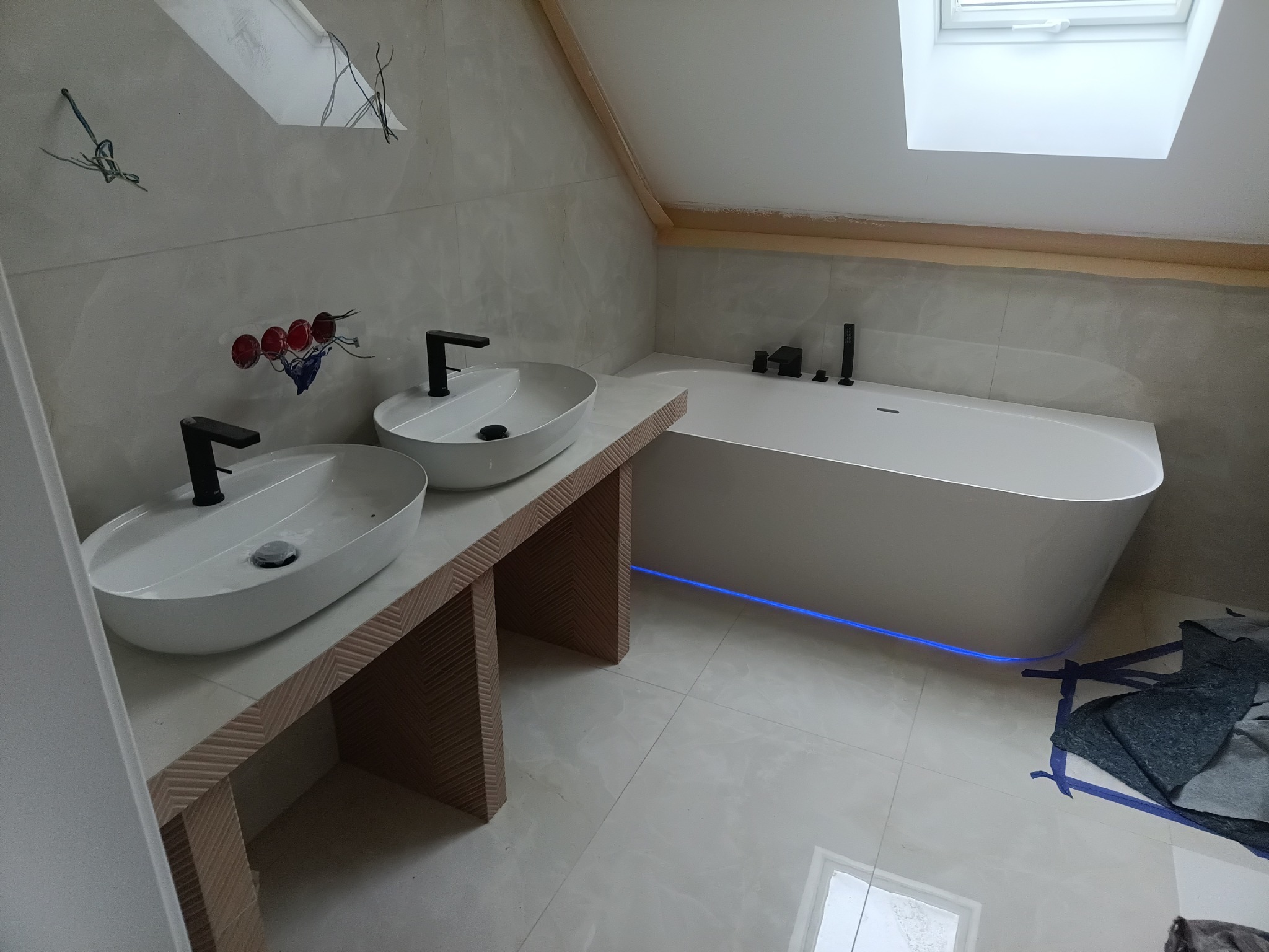 Nowoczesna łazienka na poddaszu z owalnymi umywalkami na drewnianym blacie i wolnostojącą wanną z podświetleniem LED. Białe płytki i okno dachowe.