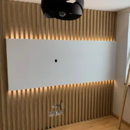 Ściana z drewnianych lameli z podświetleniem LED, biały panel centralny z otworem, widoczne gniazdka elektryczne i czarna, nowoczesna lampa sufitowa.