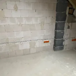 Surowy stan poddasza z widoczną instalacją elektryczną: kable w peszlach i pomarańczowe puszki podtynkowe na ścianie z białych bloczków.