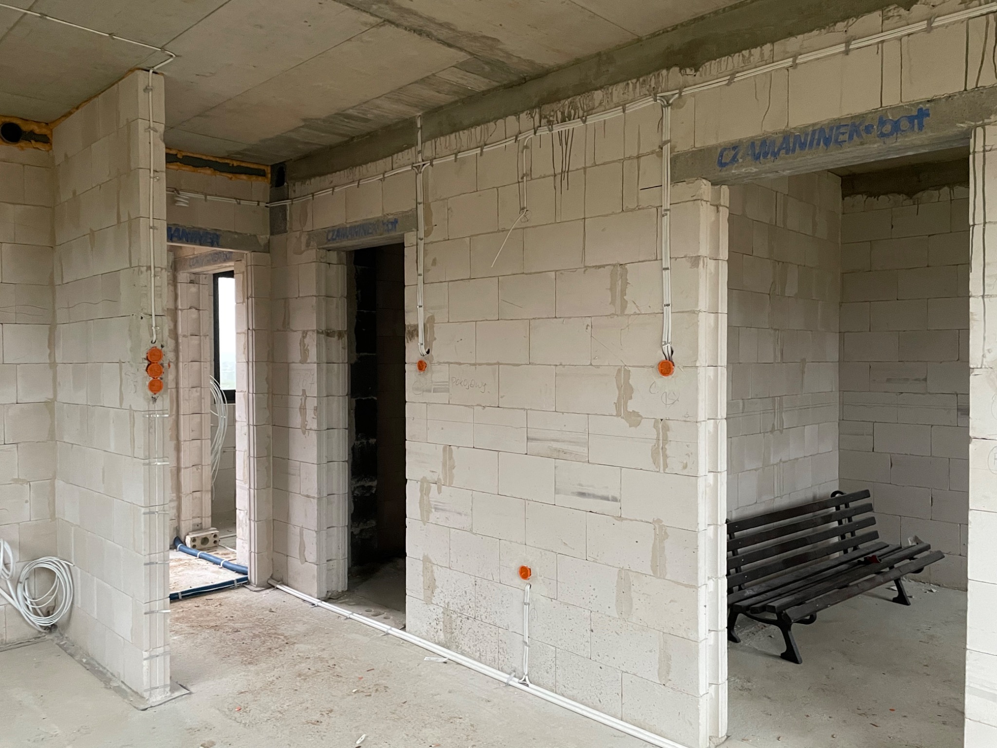 Surowy stan wnętrza budynku w trakcie prac, widoczne białe ściany z betonu komórkowego, wystające przewody elektryczne w białych listwach, puszki elektryczne, napis 'CZ.WANINEK-bot' nad otworem...