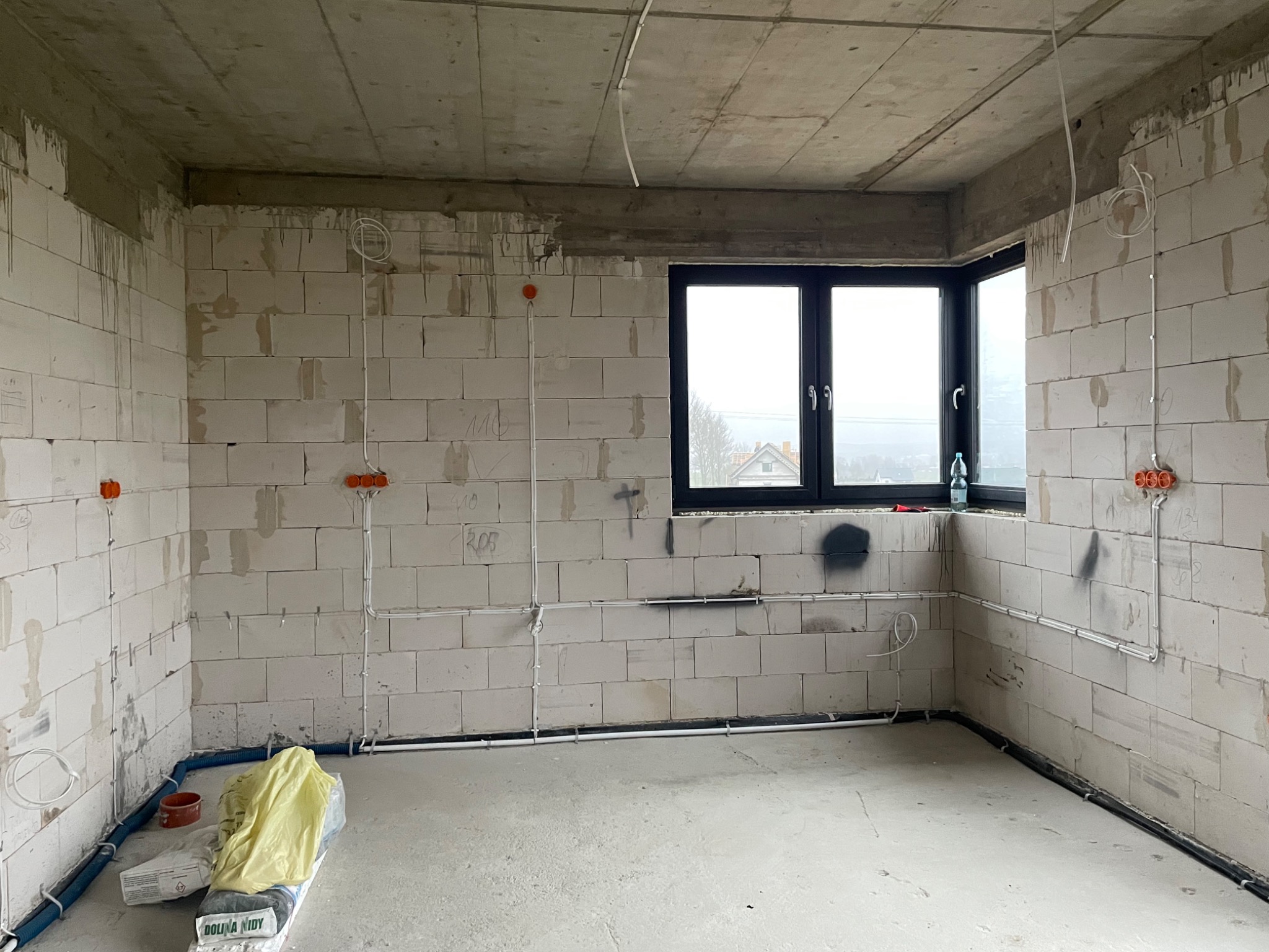 Surowy stan wnętrza z widocznymi przewodami elektrycznymi poprowadzonymi po ścianach z betonu komórkowego, puszki instalacyjne w kolorze pomarańczowym, niebieskie rury karbowane przy podłodze, okno...