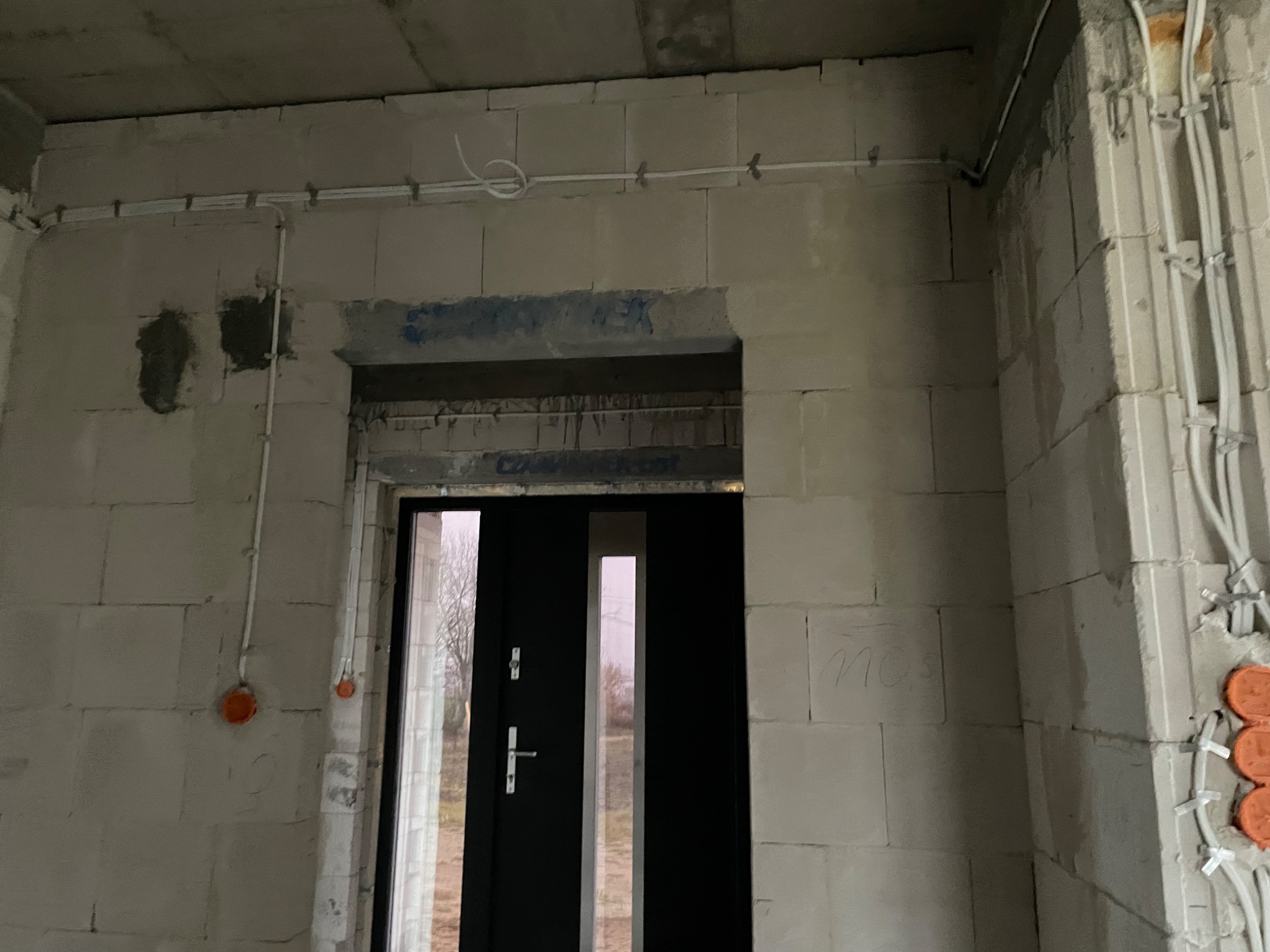 Instalacja elektryczna w surowym budynku: białe przewody prowadzone po ścianach z bloczków, widoczne puszki instalacyjne i nowa czarna stolarka drzwiowa z przeszkleniami.