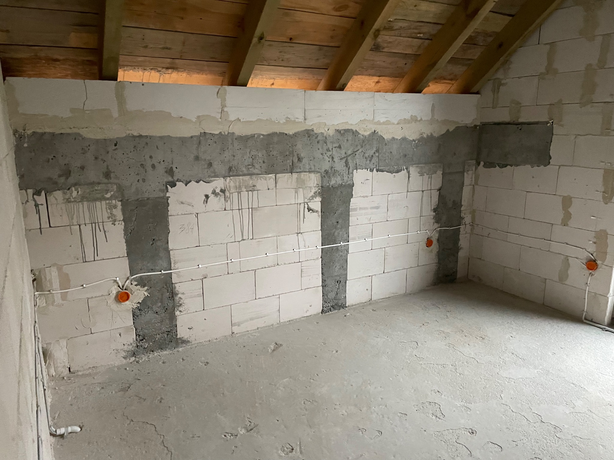 Surowy stan poddasza z widocznymi przewodami elektrycznymi w białych rurkach, pomarańczowe puszki instalacyjne na ścianach z betonu komórkowego, betonowa wylewka na podłodze i drewniany strop.