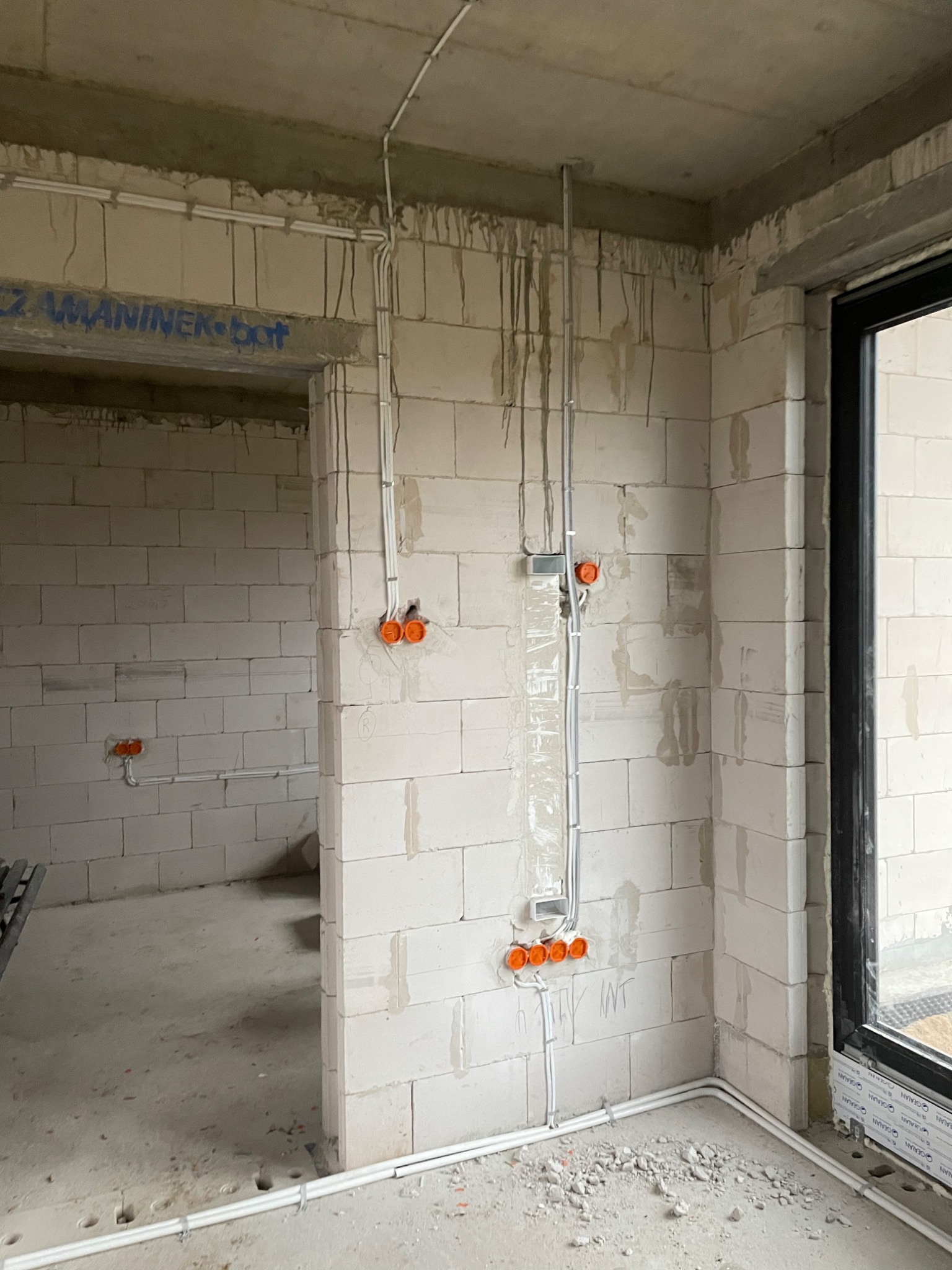 Surowy stan wnętrza z widocznymi przewodami elektrycznymi w peszlach i pomarańczowymi puszkami instalacyjnymi na ścianach z betonu komórkowego, widoczne okno z czarną ramą i instalacja podtynkowa...