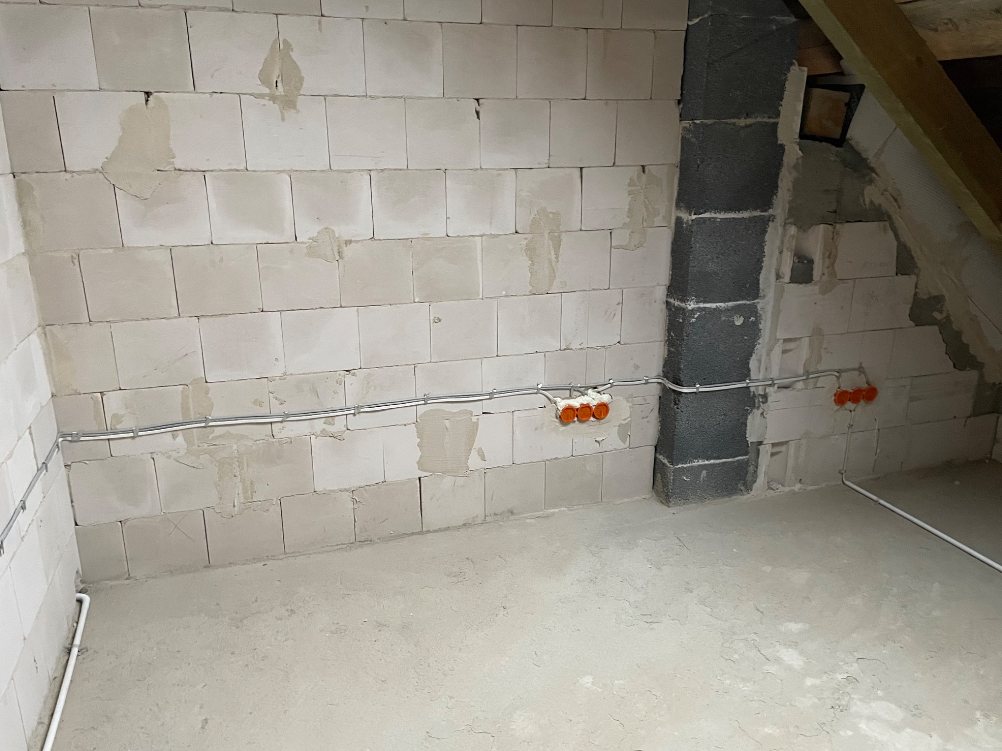 Surowy stan poddasza z widoczną instalacją elektryczną: kable w peszlach i pomarańczowe puszki podtynkowe na ścianie z białych bloczków.