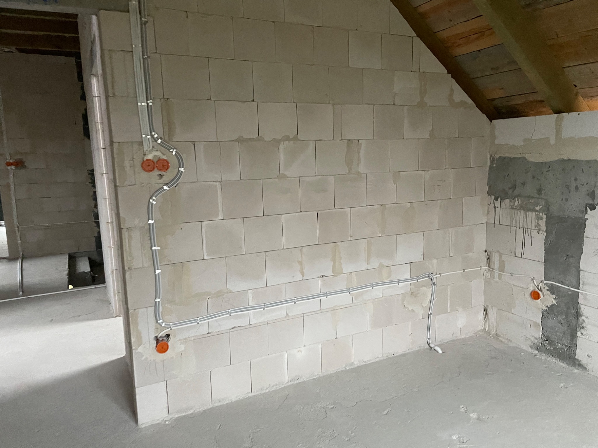 Surowe wnętrze w trakcie prac wykończeniowych: widoczne poprowadzone przewody elektryczne w peszlach na ścianie z betonu komórkowego, puszki elektryczne w kolorze pomarańczowym, surowy beton...