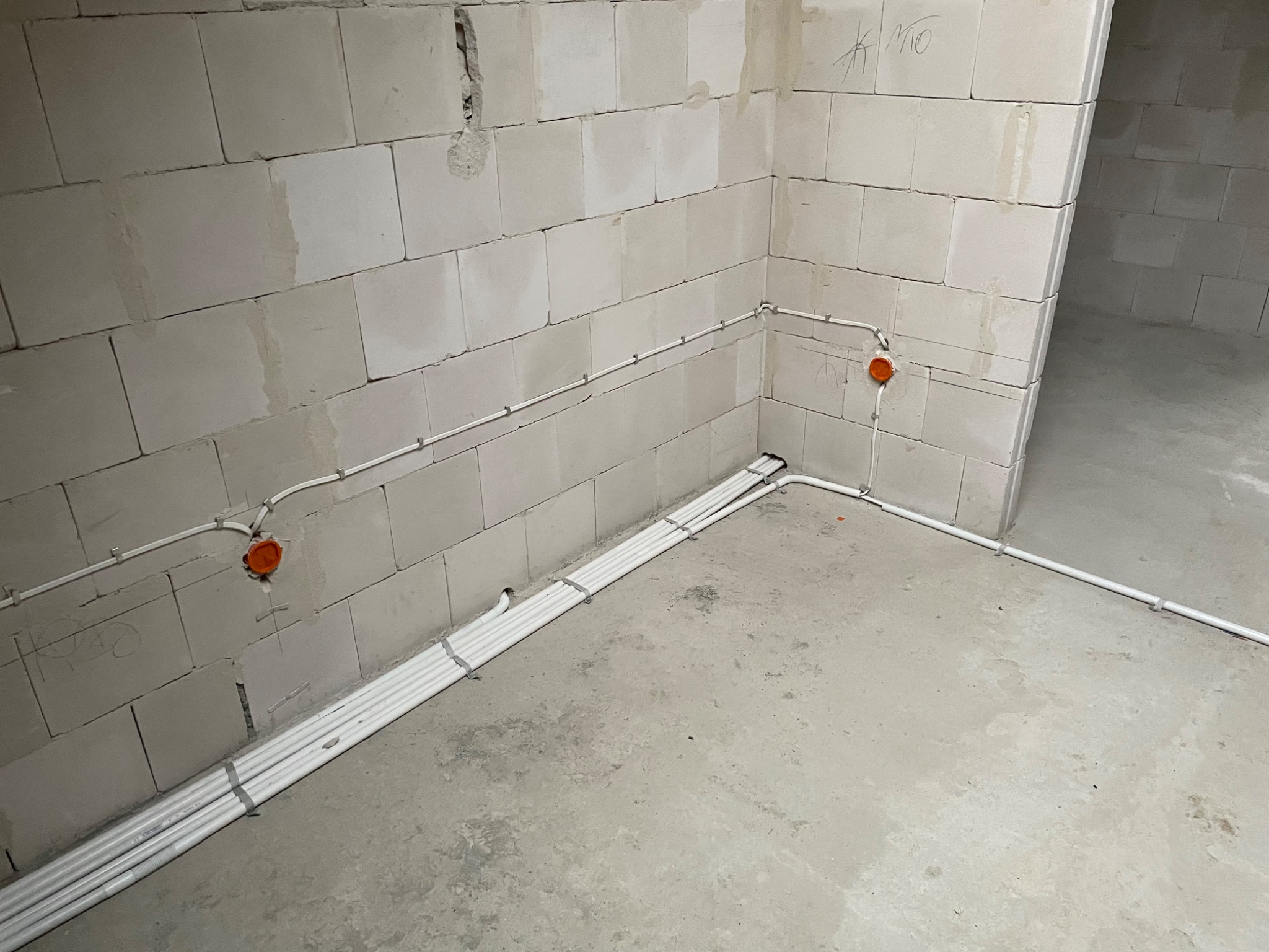 Wewnętrzne prowadzenie przewodów elektrycznych w białych rurkach, mocowanych klamrami do surowej ściany z betonu komórkowego, widoczna pomarańczowa puszka instalacyjna.