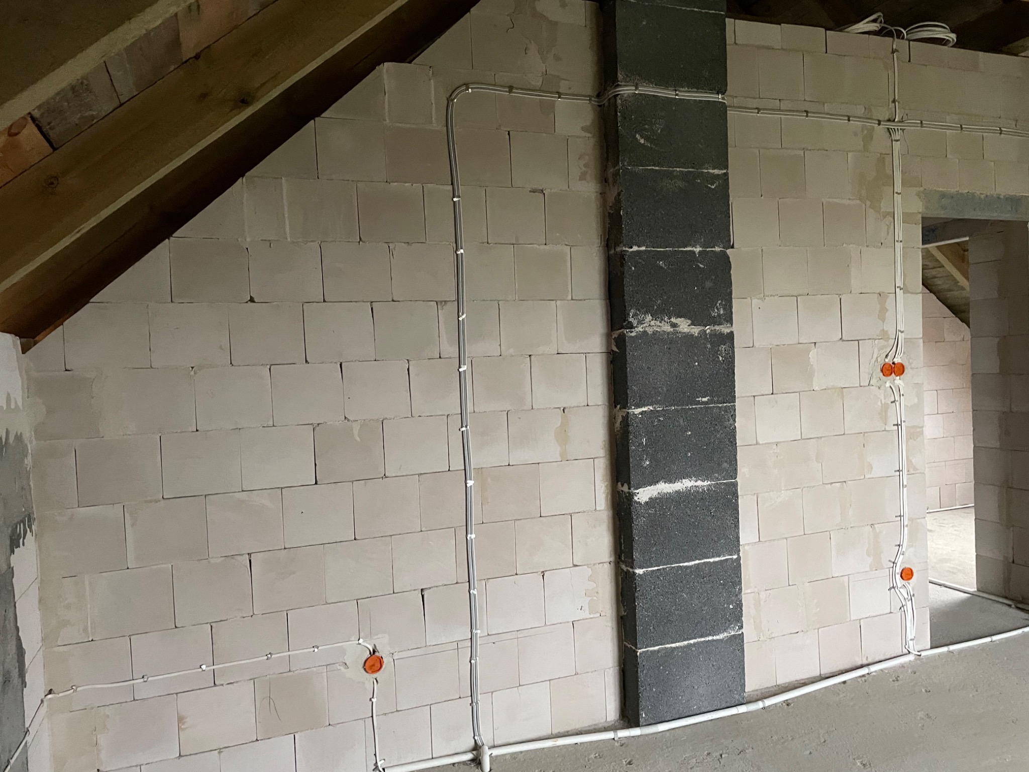 Surowy stan poddasza z instalacją elektryczną: białe przewody prowadzone po ścianach z bloczków, pomarańczowe puszki podtynkowe, fragment komina z szarej cegły.