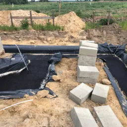Wykonanie izolacji poziomej fundamentów z użyciem czarnej folii hydroizolacyjnej i bloczków betonowych, widoczne wykopy i hałdy ziemi w tle.