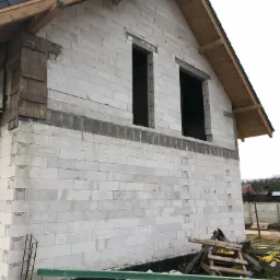 Surowa bryła domu z keramzytobetonu w trakcie budowy, widoczne otwory okienne bez ram, drewniana konstrukcja dachu i elementy szalunkowe przy oknie, teren budowy z gruzem i folią.