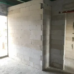 Wnętrze w trakcie budowy z widocznymi ścianami z białych bloczków, surowy beton na podłodze z gruzem, oraz widoczne elementy konstrukcji dachu.