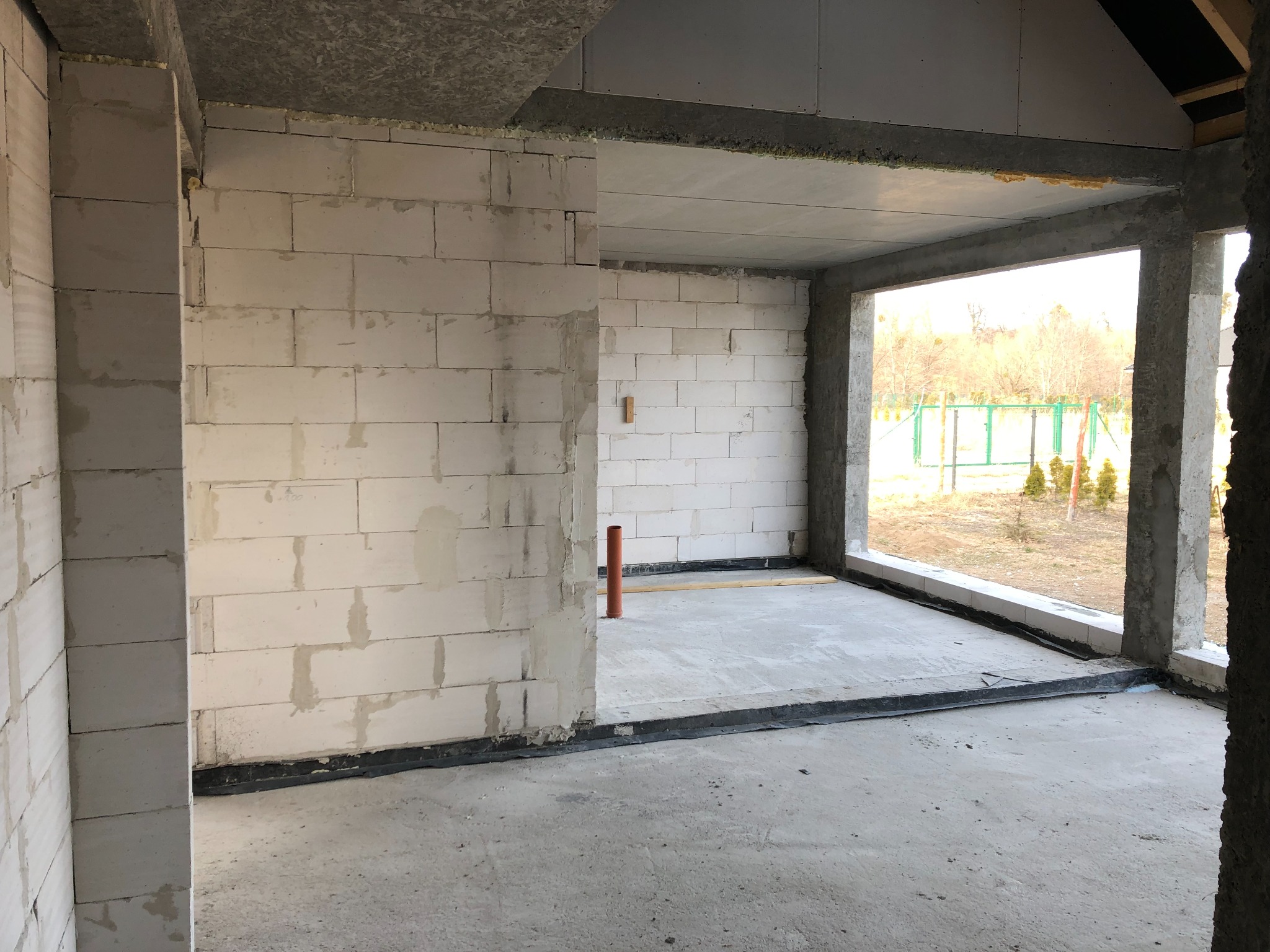 Wnętrze surowego domu z białych bloczków, widoczne ściany, surowy betonowy strop i przygotowane otwory okienne, na zewnątrz widok na ogród z zielonym ogrodzeniem.