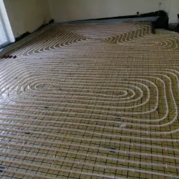 Układanie rur ogrzewania podłogowego na folii z siatką, widoczne okno i fragment ściany w surowym stanie.